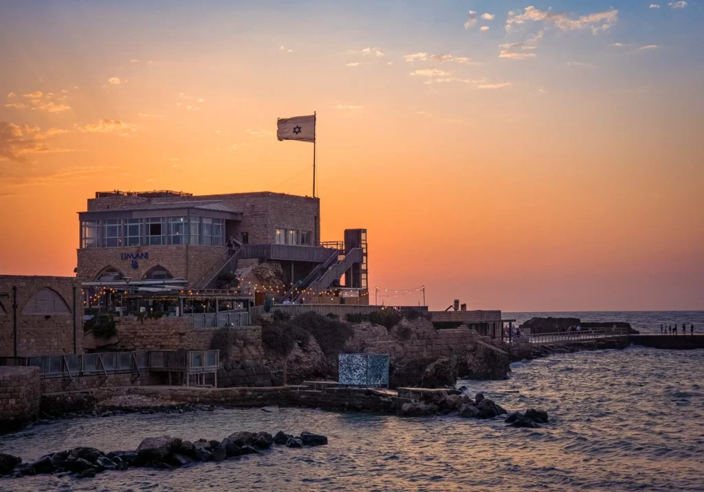 השמש שוקעת מאחורי מסעדה עם דגל ישראל מתנופף מעליה, הנמצאת במזח בנמל ההיסטורי של קיסריה, צופה אל הים התיכון עם חוף סלעי בחזית