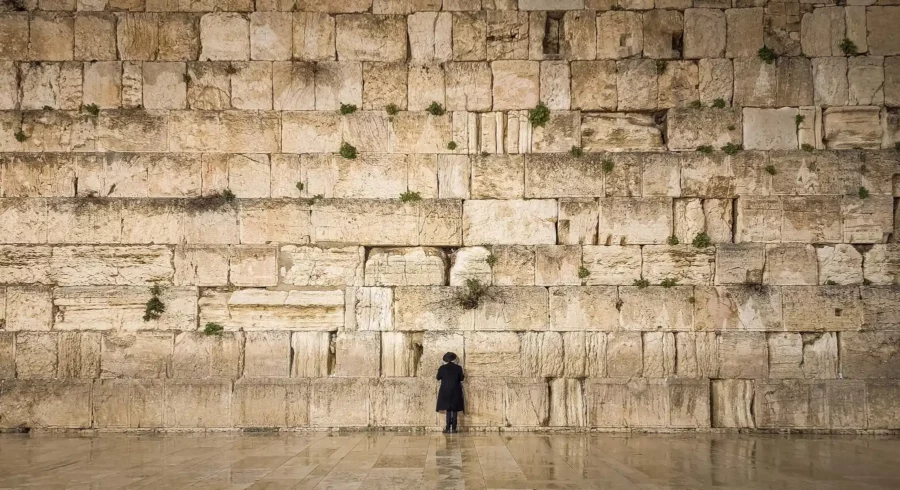 יהודי שלבוש כחרדי עומד בתפילה בהתבודדות מול הכותל המערבי בירושלים, משקף את המשמעות ההיסטורית והרוחנית של המקום הקדוש