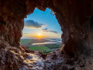 שקיעה מרהיבה שנצפית דרך קשת טבעית של מערה ברכס הכרמל, עם נוף לשמים תכולים עם עננים, שדות ירוקים, ולחוף הים