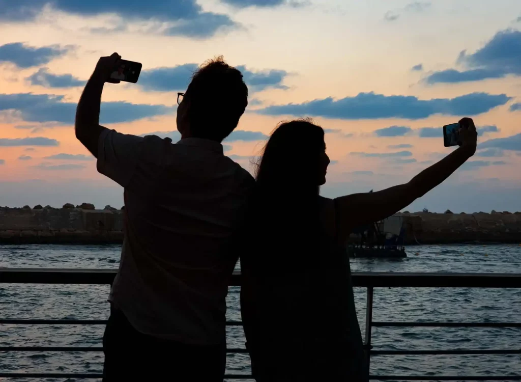 צללית של זוג מצלם סלפי עם סמארטפון על רקע שקיעה מול הים בנמל יפו במהלך סדנת צילום בסמארטפון לזוג
