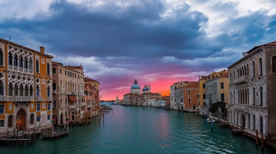 התעלה הגדולה בוונציה עטופה באור הרך של הזריחה בגווני אדום־כתום, עם בזיליקת סנטה מריה דלה סאלוטה ברקע.