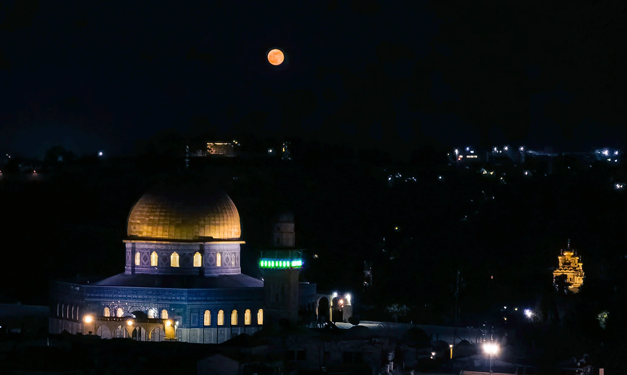 כיפת הזהב של הר הבית עם שמי לילה בהירים עם ירח מלא זוהר העולה ברקע, ואורות העיר ירושלים מנצנצים למטה.