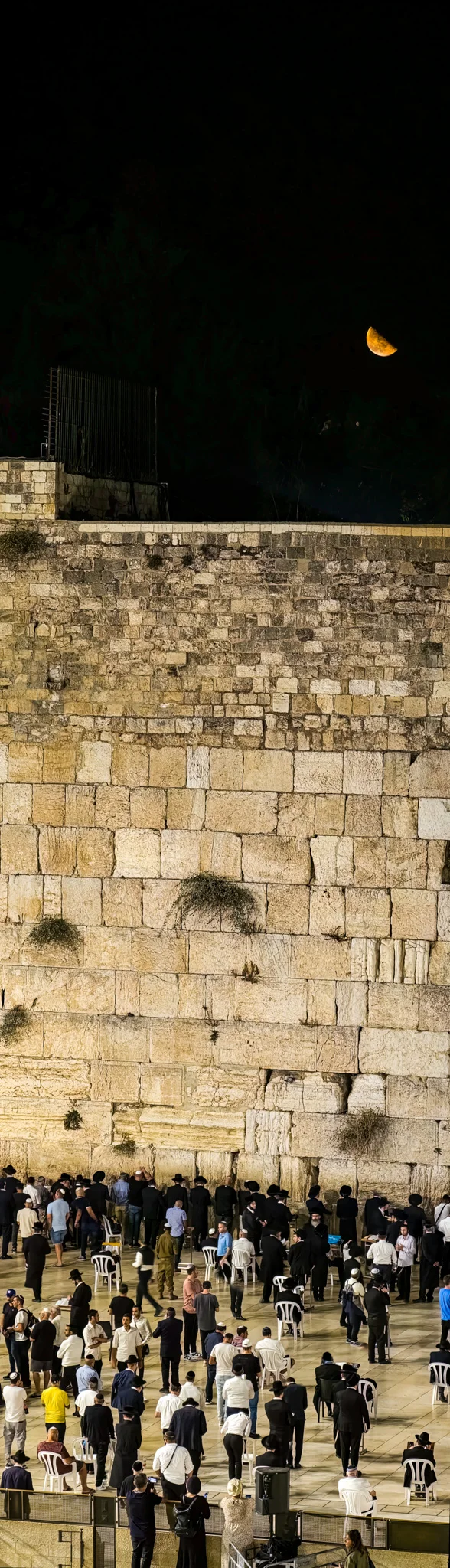 אנשים מתפללים בכותל עם סהר הירח תלוי בשמים האפלים מעל ירושלים.