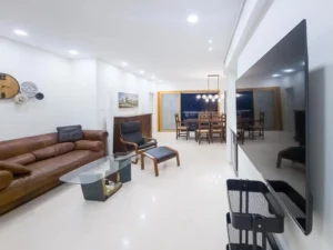 סלון מודרני עם ספה עור חומה, שולחן זכוכית, כורסה שחורה וטלוויזיה גדולה, כולל פינת אוכל ברקע עם שולחן עץ וכיסאות, שצולמה במהלך סדנת צילום בסמארטפון לצילום אדריכלות ועיצוב פנים