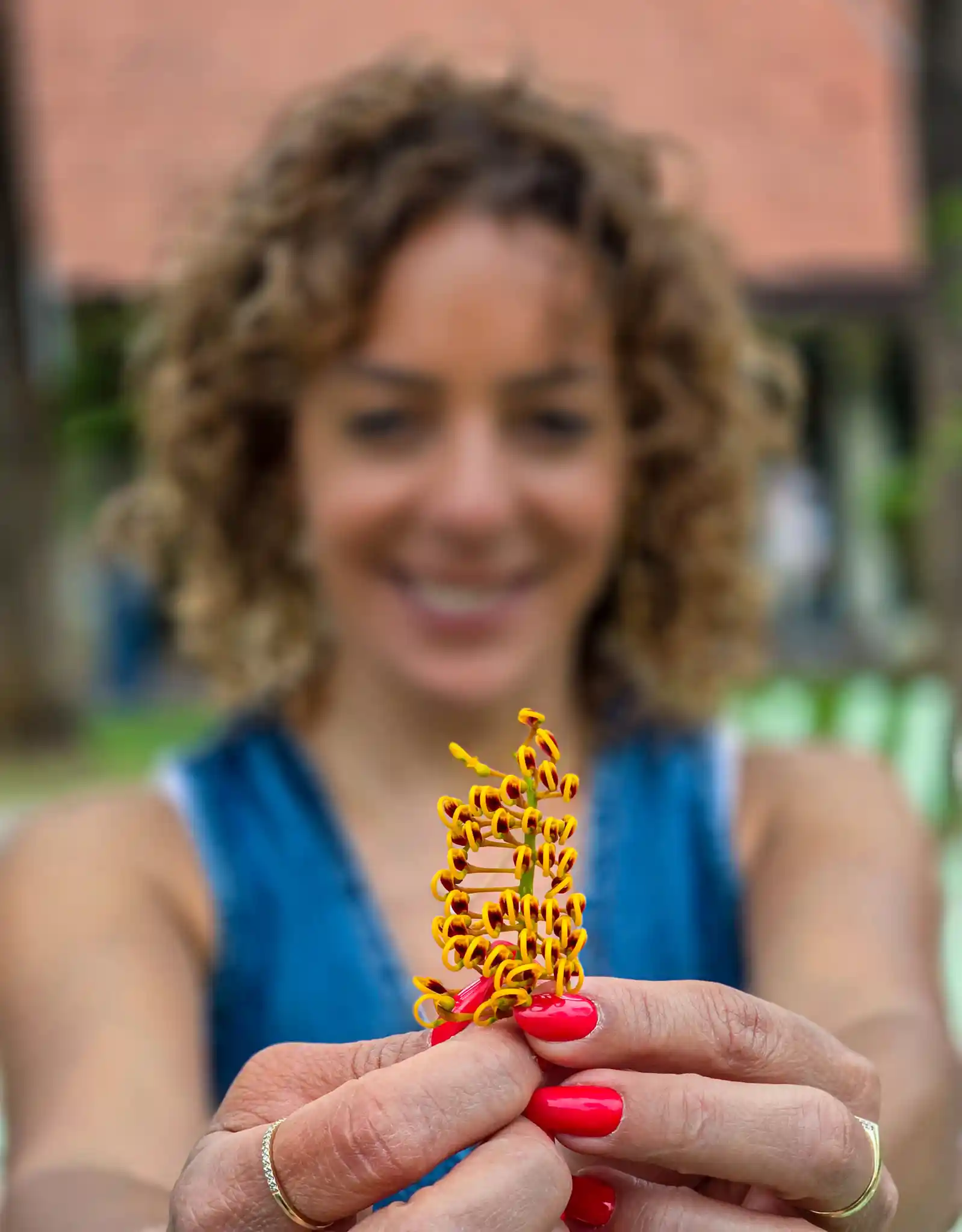 אישה מחייכת ומחזיקה בפריים הקדמי חלק צבעוני של פרח קטן עם פרטים צהובים-אדומים מסולסלים, כשידיה בפוקוס מלא והפנים ברקע מטושטשות. הצילום נעשה בגינת מלון הגושרים במהלך סדנת צילום בסמארטפון לצוות הנשי של אוניברסיטת תל חי, כהדגמה לעבודה עם עומק שדה ומיקוד ידני.