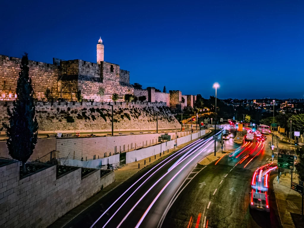 מבט לילי אל חומות העיר העתיקה בירושלים ומגדל דוד משמאל, עם פסי אור של תנועת רכבים שצולמו בצילום ארוך חשיפה בשעה הכחולה. התמונה מדגישה את הניגוד בין תאורת הרחוב החמימה לשמיים הכחולים העמוקים, כפי שנלמד במסגרת סדנת צילום בסמארטפון.