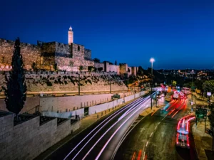 מבט לילי אל חומות העיר העתיקה בירושלים ומגדל דוד משמאל, עם פסי אור של תנועת רכבים שצולמו בצילום ארוך חשיפה בשעה הכחולה. התמונה מדגישה את הניגוד בין תאורת הרחוב החמימה לשמיים הכחולים העמוקים, כפי שנלמד במסגרת סדנת צילום בסמארטפון.