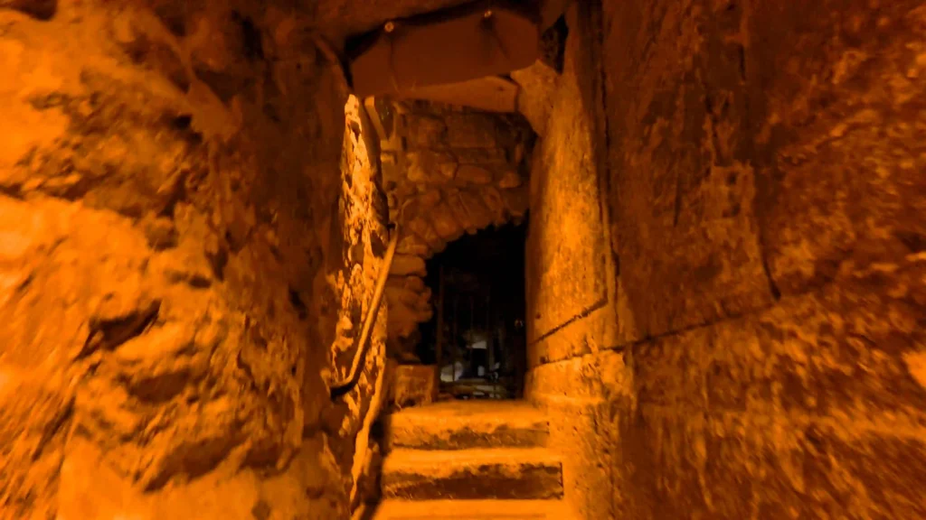 מדרגות אבן עתיקות ומעבר צר וחשוך המוארים בתאורה חמה, בירידה לעומק מנהרות הכותל המערבי בירושלים.